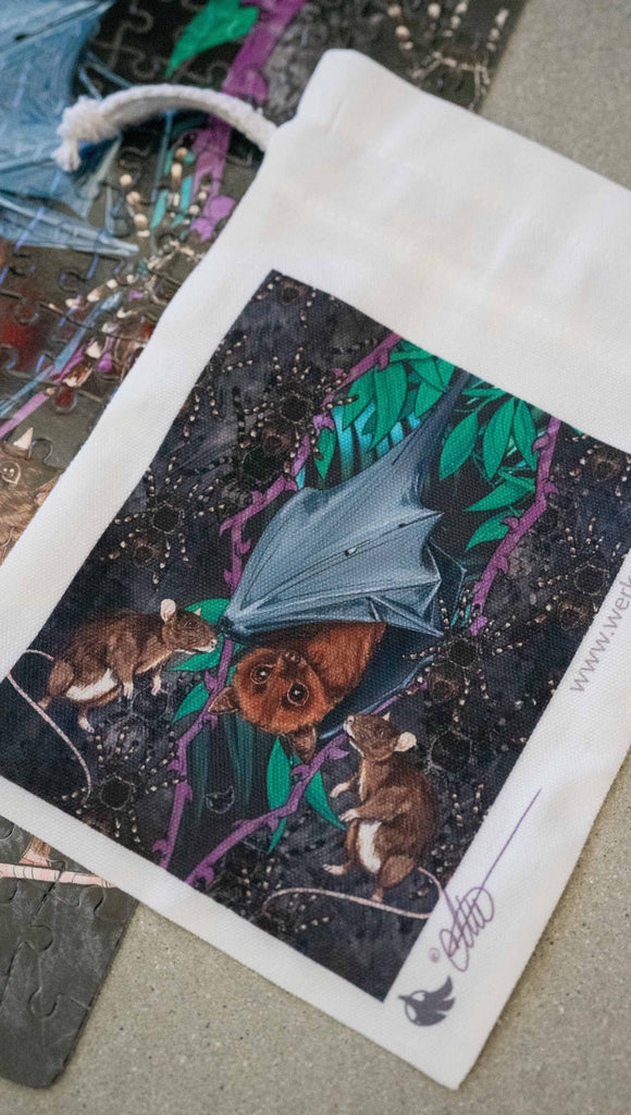 WERKSHOP Spooky Season jigsaw puzzle The artwork features an adorable fruit bat dangling upside down inside a tropical scene with a purple wreath of thorns. Under the bat, there are two rats. The background is a distressed dark gray brushstroke texture. The puzzle comes with a canvas drawcord pouch.