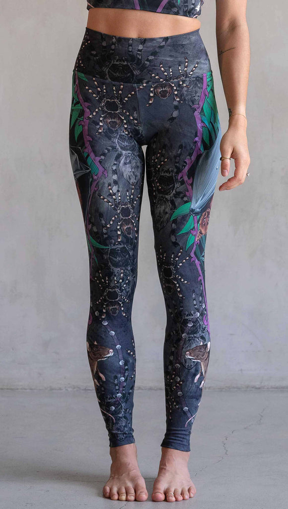 Model wearing WERKSHOP Spooky Season Set. The leggings feature an adorable fruit  bat dangling upside down inside a tropical scene with a purple wreath of thorns. under the bat, there is a rat facing forward. The background is a distressed dark gray brushstroke texture with scattered tarantulas.