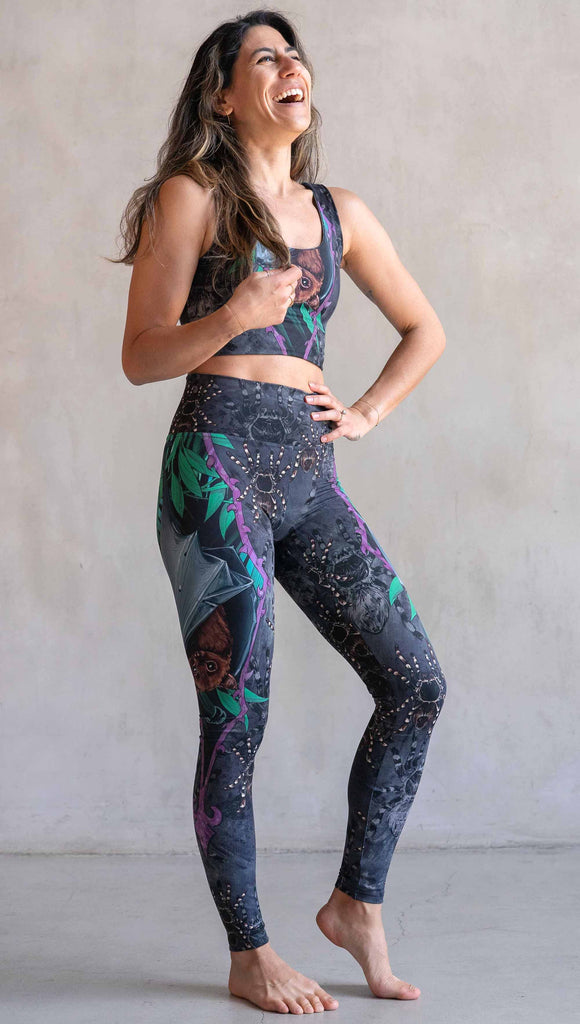 Model wearing WERKSHOP Spooky Season Set. The leggings feature an adorable fruit bat dangling upside down inside a tropical scene with a purple wreath of thorns. The background is a distressed dark gray brushstroke texture. (This version of the artwork has no rats)