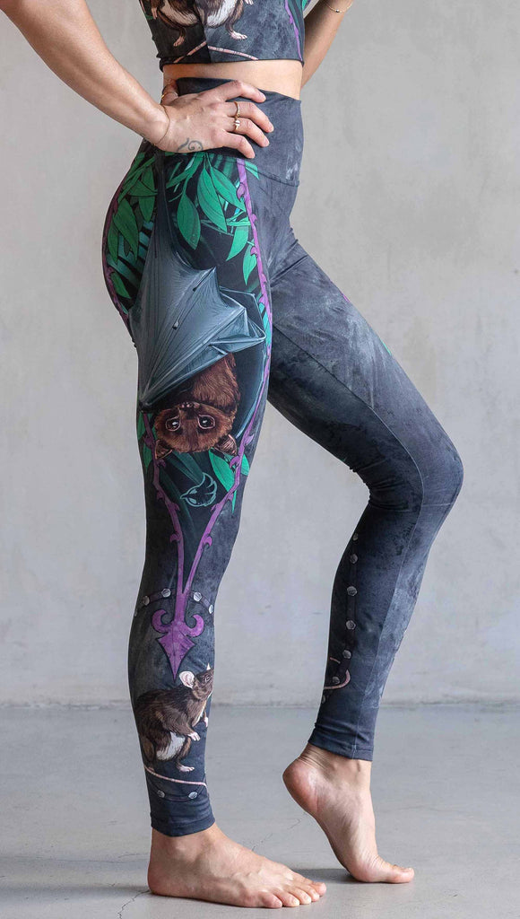 Model wearing WERKSHOP Spooky Season Set. The leggings feature an adorable fruit bat dangling upside down inside a tropical scene with a purple wreath of thorns. under the bat, there is a rat facing forward. The background is a distressed dark gray brushstroke texture. (This version of the artwork has no spiders)