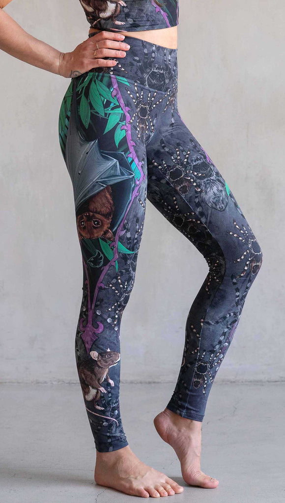 Model wearing WERKSHOP Spooky Season Set. The leggings feature an adorable fruit bat dangling upside down inside a tropical scene with a purple wreath of thorns. under the bat, there is a rat facing forward. The background is a distressed dark gray brushstroke texture with scattered tarantulas.
