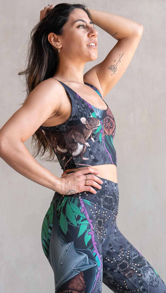 Model wearing WERKSHOP Spooky Season reversible top. One side features an adorable fruit bat dangling upside down inside a tropical scene with a purple wreath of thorns. under the bat, there is a rat facing forward. The background is a distressed dark gray brushstroke texture with scattered tarantulas. The other side is just "ghost" tarantulas.