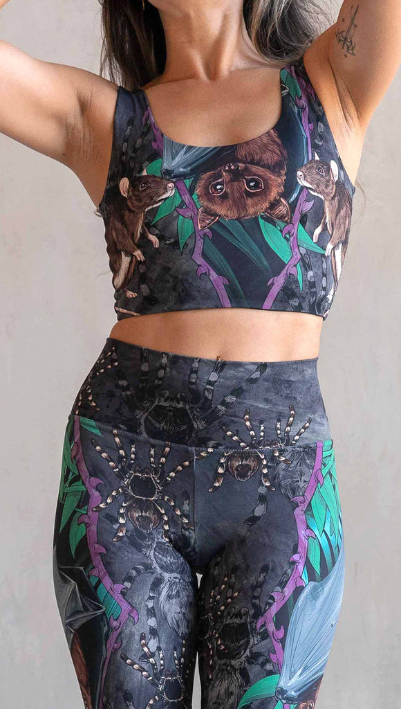 Model wearing WERKSHOP Spooky Season reversible top. One side features an adorable fruit bat dangling upside down inside a tropical scene with a purple wreath of thorns. under the bat, there is a rat facing forward. The background is a distressed dark gray brushstroke texture with scattered tarantulas. The other side is just "ghost" tarantulas.
