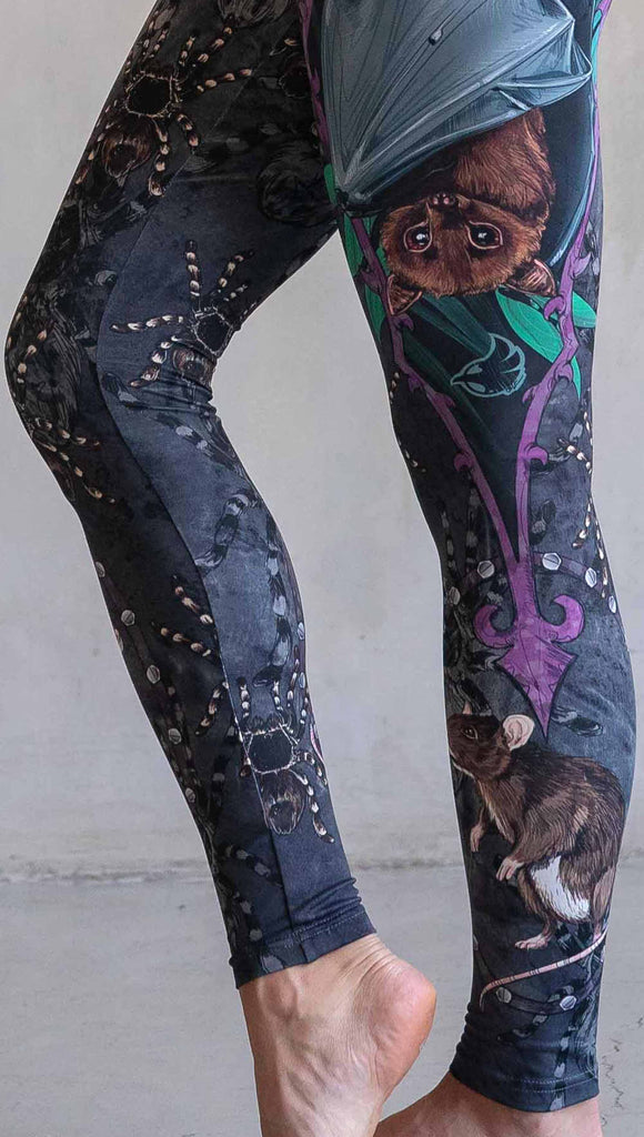 Zoomed in photo of model wearing WERKSHOP Spooky Season Set. The leggings feature an adorable fruit  bat dangling upside down inside a tropical scene with a purple wreath of thorns. under the bat, there is a rat facing forward. The background is a distressed dark gray brushstroke texture with scattered tarantulas.