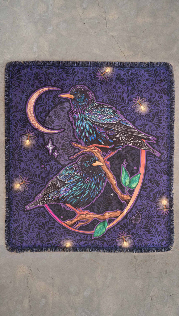 WERKHOP Starlings tapestry laid flat on a concrete floor. The tapestry is printed with original artwork by Chriztina Marie fearuring Featuring European Starlings perched on a branch near a crescent moon and fireflies. The colors are warm purples with pops of pink, gold and green