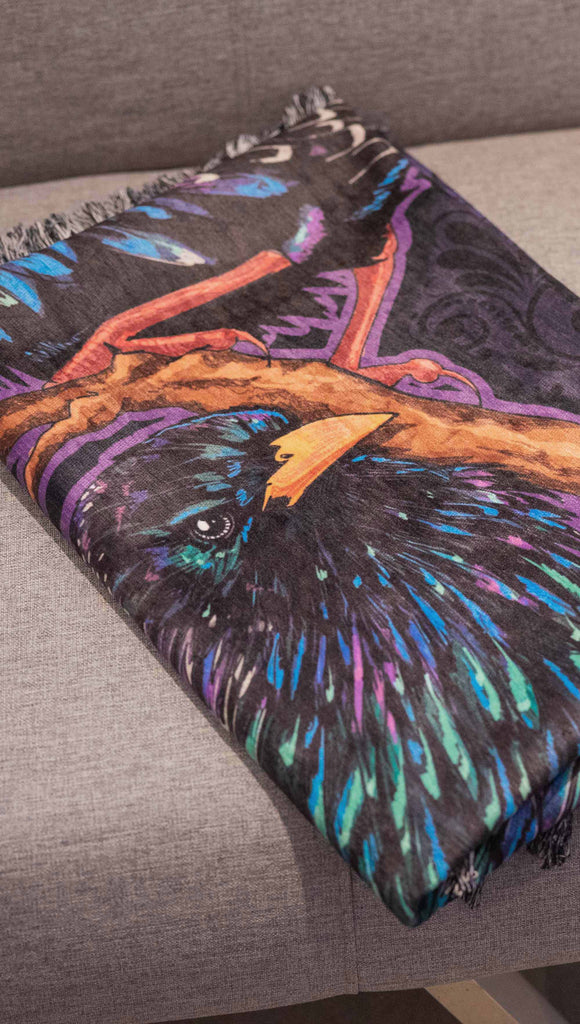 WERKSHOP Starlings tapestry neatly folded on a futon. The tapestry is printed with original artwork by Chriztina Marie fearuring Featuring European Starlings perched on a branch near a crescent moon and fireflies. The colors are warm purples with pops of pink, gold and green