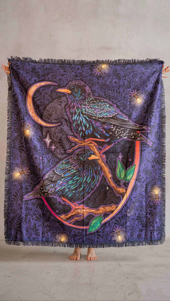 WERKSHOP Starlings tapestry draped on a couch. The tapestry is printed with original artwork by Chriztina Marie fearuring Featuring European Starlings perched on a branch near a crescent moon and fireflies. The colors are warm purples with pops of pink, gold and green.