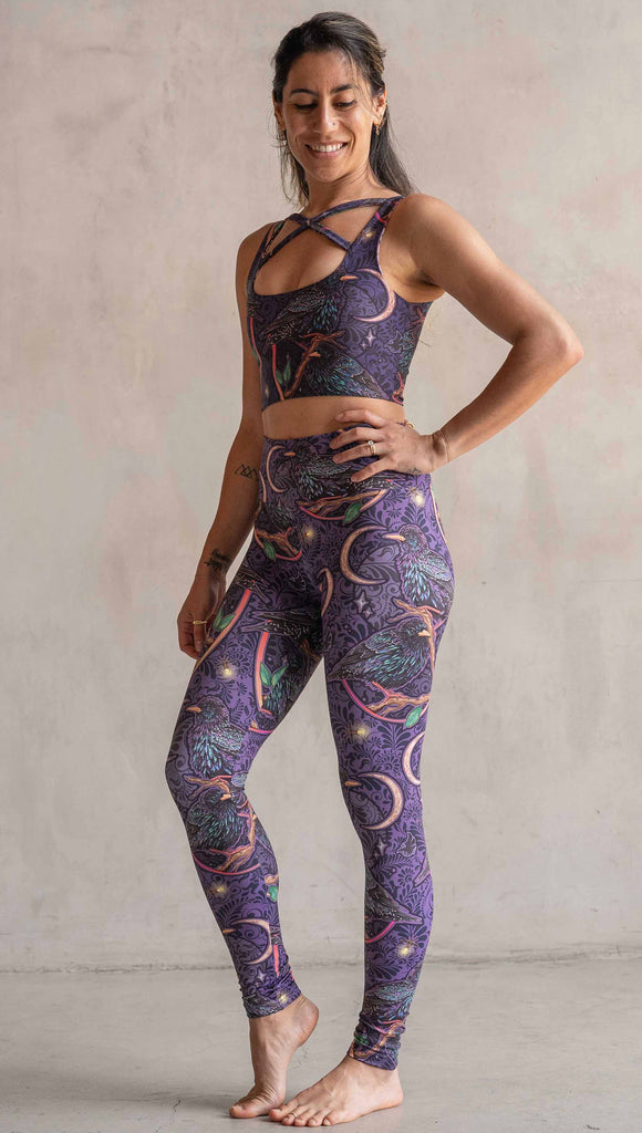 Full body side view of model wearing WERKSHOP Starlings Athleisure Leggings. The fabric is printed with original artwork by our Female Founder, Chriztina Marie. Featuring European Starlings perched on a branch near a crescent moon and fireflies. The colors are warm purples with pops of pink, gold and green.