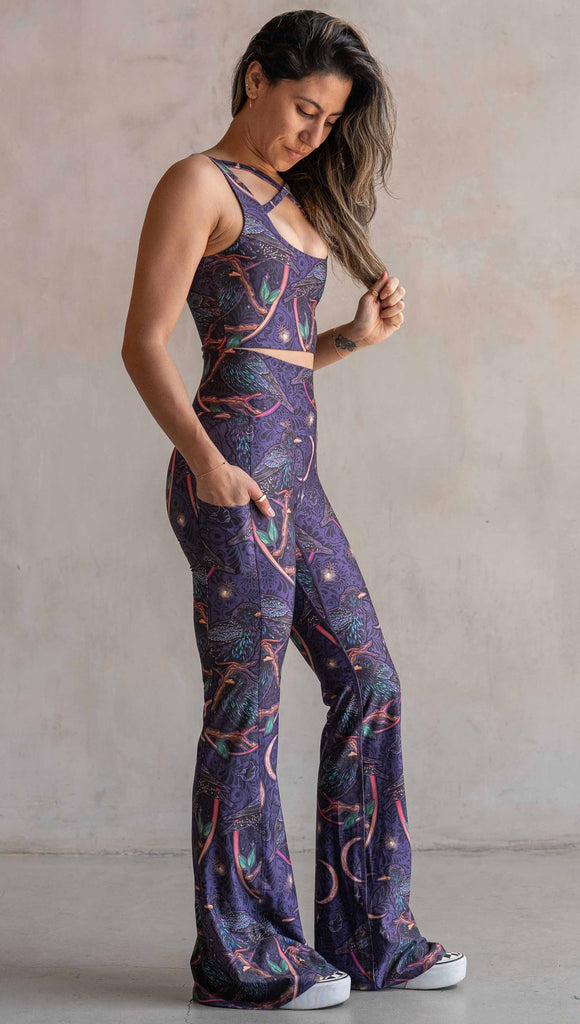 Full body side view of model wearing WERKSHOP Starlings EnviSoft bell bottom pants with pockets. The fabric is printed with original artwork by our Female Founder, Chriztina Marie. Featuring European Starlings perched on a branch near a crescent moon and fireflies. The colors are warm purples with pops of pink, gold and green.