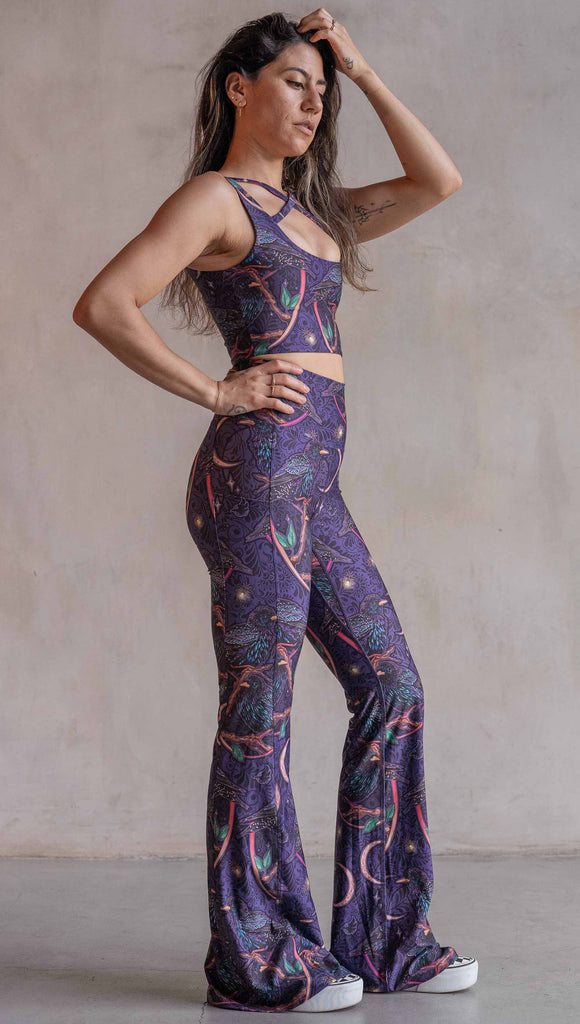 Fulll body side view of model wearing WERKSHOP Starlings EnviSoft bell bottom pants with pockets. The fabric is printed with original artwork by our Female Founder, Chriztina Marie. Featuring European Starlings perched on a branch near a crescent moon and fireflies. The colors are warm purples with pops of pink, gold and green.