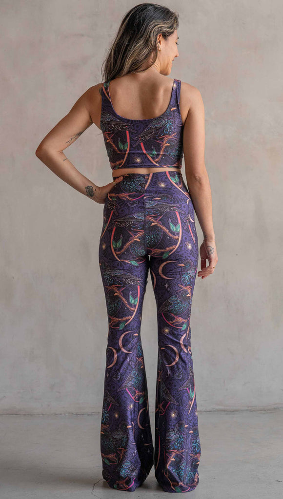 Full body back view of model wearing WERKSHOP Starlings EnviSoft bell bottom pants with pockets. The fabric is printed with original artwork by our Female Founder, Chriztina Marie. Featuring European Starlings perched on a branch near a crescent moon and fireflies. The colors are warm purples with pops of pink, gold and green.