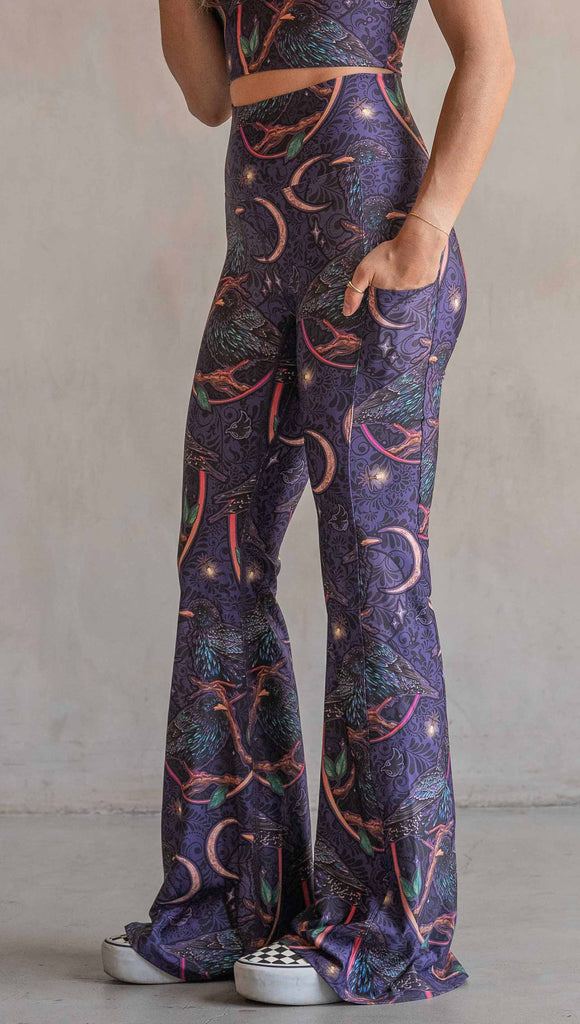 Side view of model wearing WERKSHOP Starlings EnviSoft bell bottom pants with pockets. The fabric is printed with original artwork by our Female Founder, Chriztina Marie. Featuring European Starlings perched on a branch near a crescent moon and fireflies. The colors are warm purples with pops of pink, gold and green.