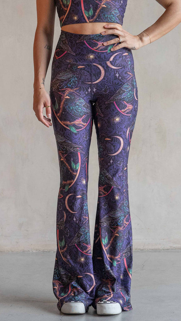 Front view of model wearing WERKSHOP Starlings EnviSoft bell bottom pants with pockets. The fabric is printed with original artwork by our Female Founder, Chriztina Marie. Featuring European Starlings perched on a branch near a crescent moon and fireflies. The colors are warm purples with pops of pink, gold and green.