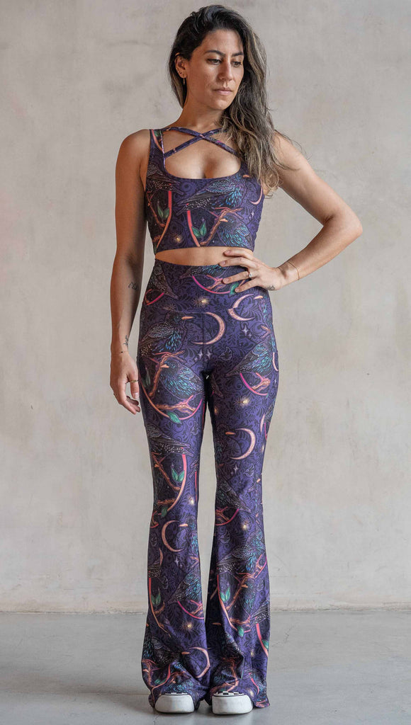 Full body front view of model wearing WERKSHOP Starlings EnviSoft bell bottom pants with pockets. The fabric is printed with original artwork by our Female Founder, Chriztina Marie. Featuring European Starlings perched on a branch near a crescent moon and fireflies. The colors are warm purples with pops of pink, gold and green.
