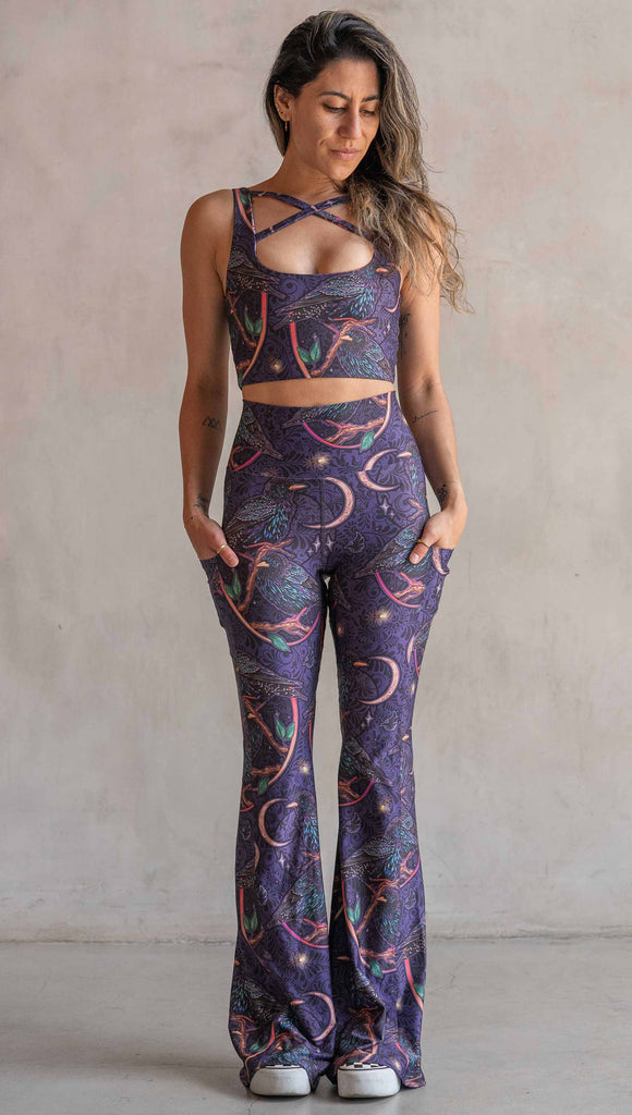 Full body front view of model wearing WERKSHOP Starlings EnviSoft bell bottom pants with pockets. The fabric is printed with original artwork by our Female Founder, Chriztina Marie. Featuring European Starlings perched on a branch near a crescent moon and fireflies. The colors are warm purples with pops of pink, gold and green.