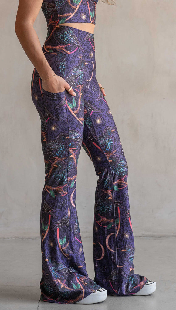 Side view of model wearing WERKSHOP Starlings EnviSoft bell bottom pants with pockets. The fabric is printed with original artwork by our Female Founder, Chriztina Marie. Featuring European Starlings perched on a branch near a crescent moon and fireflies. The colors are warm purples with pops of pink, gold and green.
