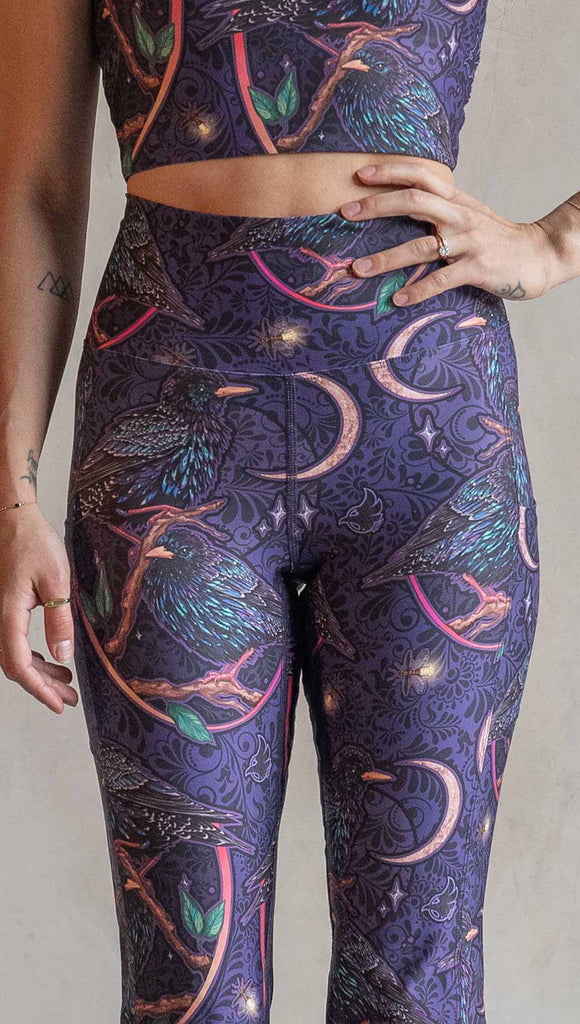 Zoomed in view of model wearing WERKSHOP Starlings EnviSoft bell bottom pants with pockets. The fabric is printed with original artwork by our Female Founder, Chriztina Marie. Featuring European Starlings perched on a branch near a crescent moon and fireflies. The colors are warm purples with pops of pink, gold and green.