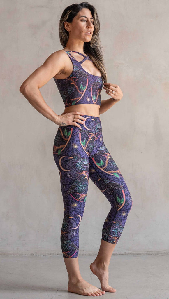 Full body side view of model wearing WERKSHOP Starlings Triathlon Capri length leggings. The fabric is printed with original artwork by our Female Founder, Chriztina Marie. Featuring European Starlings perched on a branch near a crescent moon and fireflies. The colors are warm purples with pops of pink, gold and green.