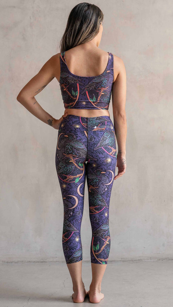 Full body back view of model wearing WERKSHOP Starlings Triathlon Capri length leggings. The fabric is printed with original artwork by our Female Founder, Chriztina Marie. Featuring European Starlings perched on a branch near a crescent moon and fireflies. The colors are warm purples with pops of pink, gold and green.