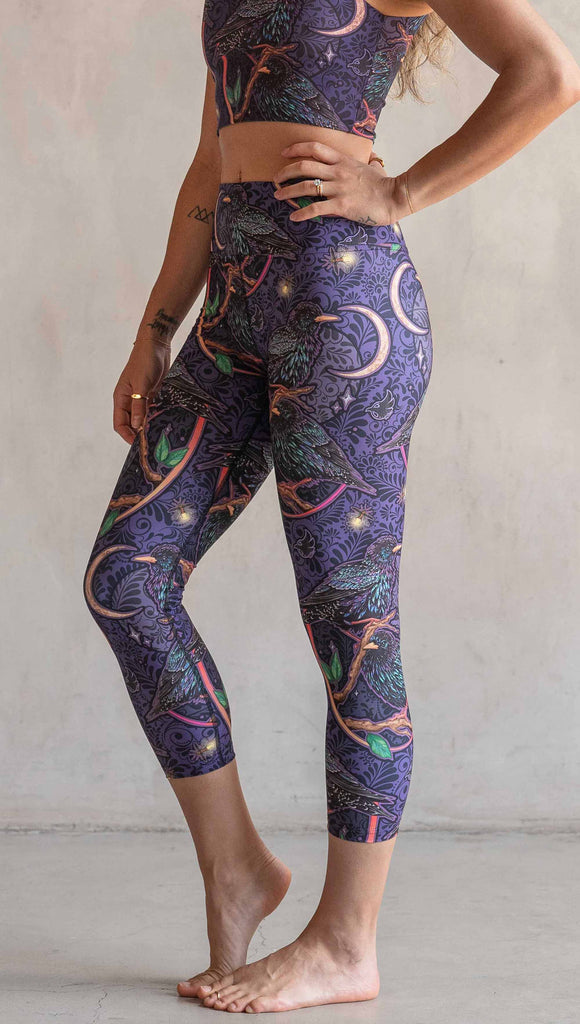 Side view of model wearing WERKSHOP Starlings Triathlon Capri length leggings. The fabric is printed with original artwork by our Female Founder, Chriztina Marie. Featuring European Starlings perched on a branch near a crescent moon and fireflies. The colors are warm purples with pops of pink, gold and green.