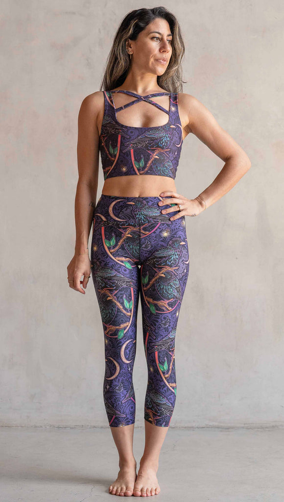 Full body front view of model wearing WERKSHOP Starlings Triathlon Capri length leggings. The fabric is printed with original artwork by our Female Founder, Chriztina Marie. Featuring European Starlings perched on a branch near a crescent moon and fireflies. The colors are warm purples with pops of pink, gold and green.