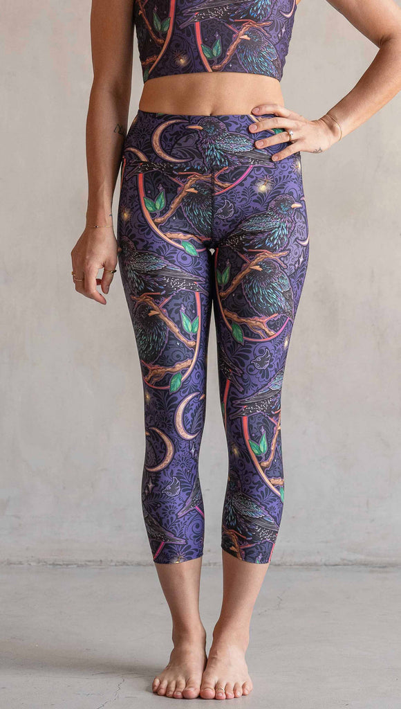 Front view of model wearing WERKSHOP Starlings Triathlon Capri length leggings. The fabric is printed with original artwork by our Female Founder, Chriztina Marie. Featuring European Starlings perched on a branch near a crescent moon and fireflies. The colors are warm purples with pops of pink, gold and green.