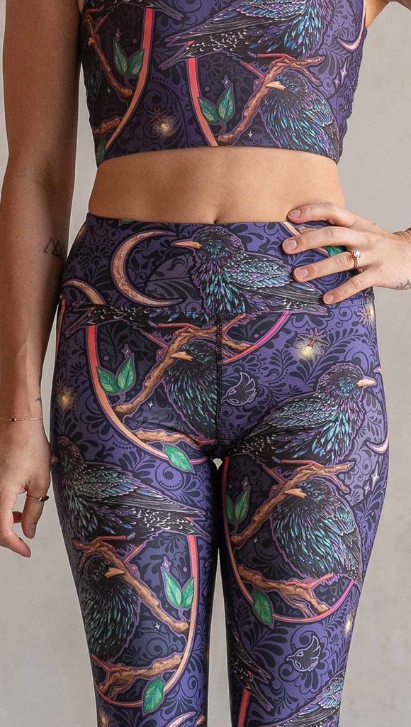 Zoomed in image of model wearing WERKSHOP Starlings Triathlon Capri length leggings. The fabric is printed with original artwork by our Female Founder, Chriztina Marie. Featuring European Starlings perched on a branch near a crescent moon and fireflies. The colors are warm purples with pops of pink, gold and green.