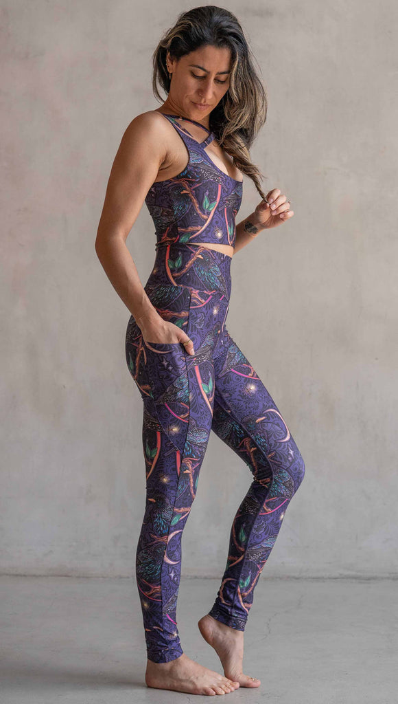 Full body side view of model wearing WERKSHOP Starlings EnviSoft leggings with pockets. The fabric is printed with original artwork by our Female Founder, Chriztina Marie. Featuring European Starlings perched on a branch near a crescent moon and fireflies. The colors are warm purples with pops of pink, gold and green.