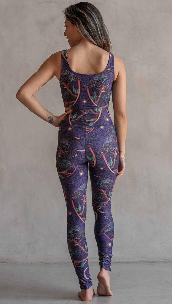 Full body back view ofmodel wearing WERKSHOP Starlings EnviSoft leggings with pockets. The fabric is printed with original artwork by our Female Founder, Chriztina Marie. Featuring European Starlings perched on a branch near a crescent moon and fireflies. The colors are warm purples with pops of pink, gold and green.