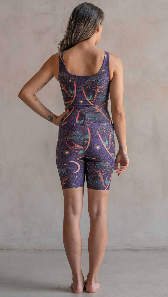 Back view of model wearing WERKSHOP Starlings EnviSoft bicycle length shorts with pockets. The fabric is printed with original artwork by our Female Founder, Chriztina Marie. Featuring European Starlings perched on a branch near a crescent moon and fireflies. The colors are warm purples with pops of pink, gold and green.