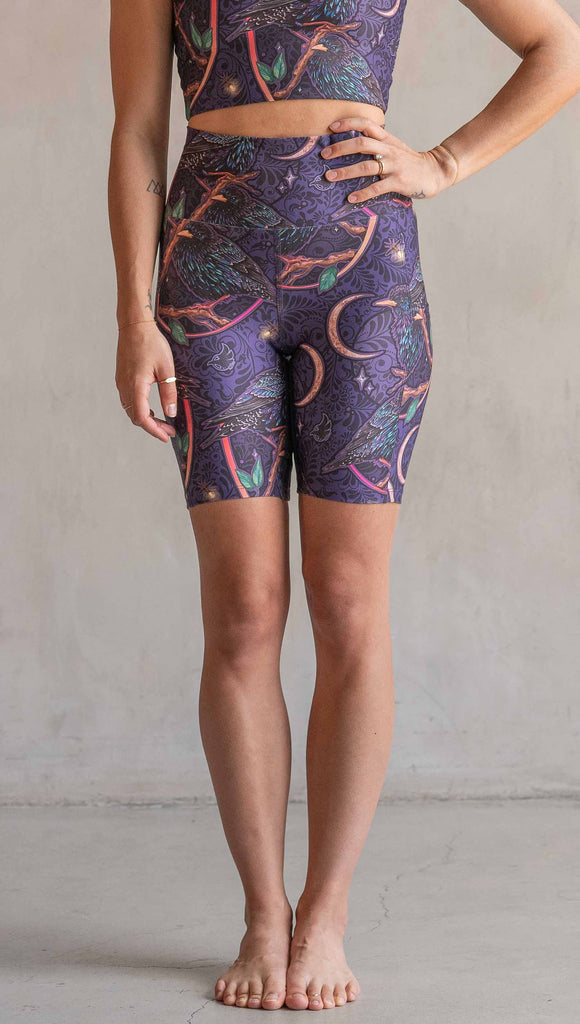 Front view of model wearing WERKSHOP Starlings EnviSoft bicycle length shorts with pockets. The fabric is printed with original artwork by our Female Founder, Chriztina Marie. Featuring European Starlings perched on a branch near a crescent moon and fireflies. The colors are warm purples with pops of pink, gold and green.