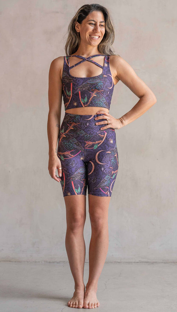 Full body front view of model wearing WERKSHOP Starlings EnviSoft bicycle length shorts with pockets. The fabric is printed with original artwork by our Female Founder, Chriztina Marie. Featuring European Starlings perched on a branch near a crescent moon and fireflies. The colors are warm purples with pops of pink, gold and green.