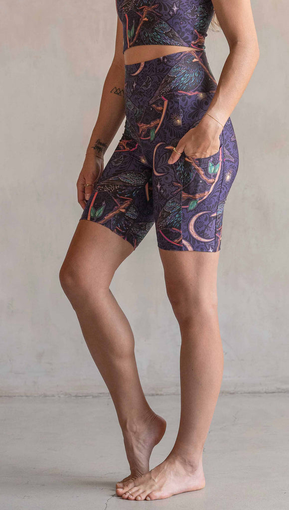 Side view of model wearing WERKSHOP Starlings EnviSoft bicycle length shorts with pockets. The fabric is printed with original artwork by our Female Founder, Chriztina Marie. Featuring European Starlings perched on a branch near a crescent moon and fireflies. The colors are warm purples with pops of pink, gold and green.
