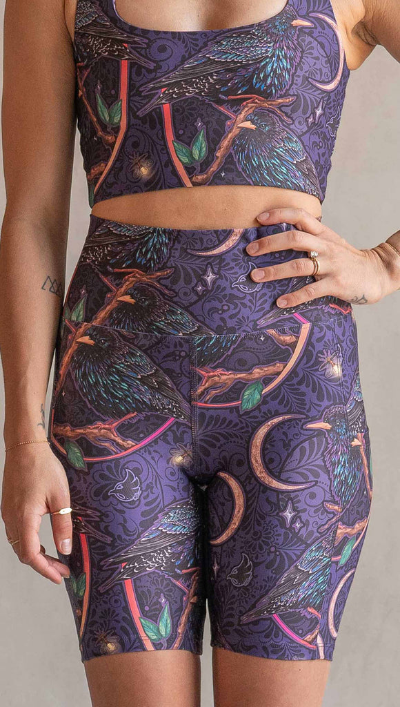 Zoomed in front view of model wearing WERKSHOP Starlings EnviSoft bicycle length shorts with pockets. The fabric is printed with original artwork by our Female Founder, Chriztina Marie. Featuring European Starlings perched on a branch near a crescent moon and fireflies. The colors are warm purples with pops of pink, gold and green.