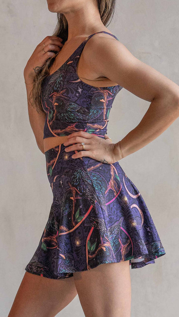 Side view of model wearing WERKSHOP Starlings EnviSoft Active Skirt with hidden shorts and pockets. The fabric is printed with original artwork by our Female Founder, Chriztina Marie. Featuring European Starlings perched on a branch near a crescent moon and fireflies. The colors are warm purples with pops of pink, gold and green.