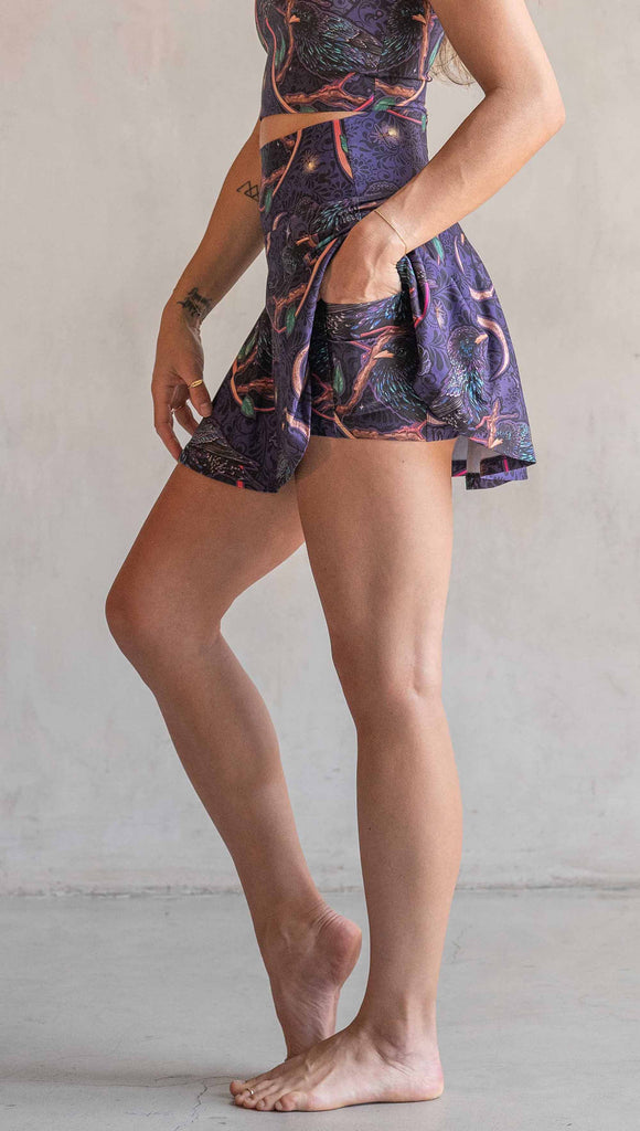 Side view of model wearing WERKSHOP Starlings EnviSoft Active Skirt with hidden shorts and pockets. The fabric is printed with original artwork by our Female Founder, Chriztina Marie. Featuring European Starlings perched on a branch near a crescent moon and fireflies. The colors are warm purples with pops of pink, gold and green.