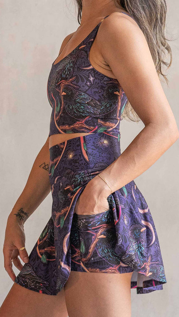 Zoomed in side view of model wearing WERKSHOP Starlings EnviSoft Active Skirt with hidden shorts and pockets. The fabric is printed with original artwork by our Female Founder, Chriztina Marie. Featuring European Starlings perched on a branch near a crescent moon and fireflies. The colors are warm purples with pops of pink, gold and green.