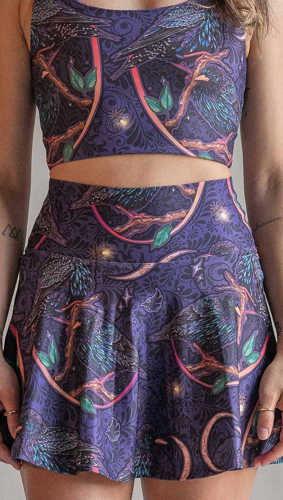 Zoomed in front view of model wearing WERKSHOP Starlings EnviSoft Active Skirt with hidden shorts and pockets. The fabric is printed with original artwork by our Female Founder, Chriztina Marie. Featuring European Starlings perched on a branch near a crescent moon and fireflies. The colors are warm purples with pops of pink, gold and green.