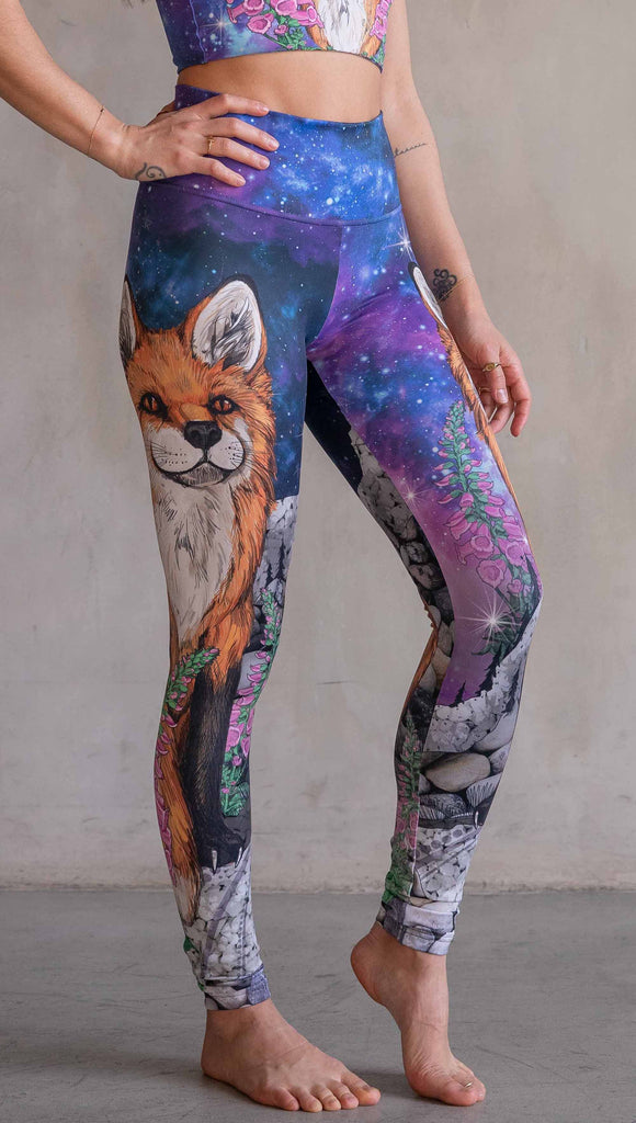 Girl wearing WERKSHOP X Save A Fox Athleisure Leggings. The artwork on the leggings features a coy red fox looking upward while perched on a rock and framed with foxglove flowers. The background for the artwork features a galactic sky with swirls of purple and blue and sparkling stars.