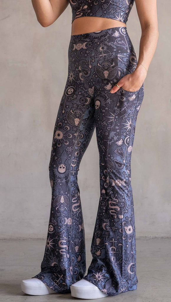 Model wearing WERKSHOP Tarot Bell Bottoms. They are high waist and feature pockets on both legs. The blue/tarot artwork has skulls, snakes, moons and the names of multiple popular tarot cards like "Strength, Lovers, Death and The Hanged Man”. Our model is 5’6” and this images shows her wearing the bells barefoot. The hem of the pants are hitting the floor.