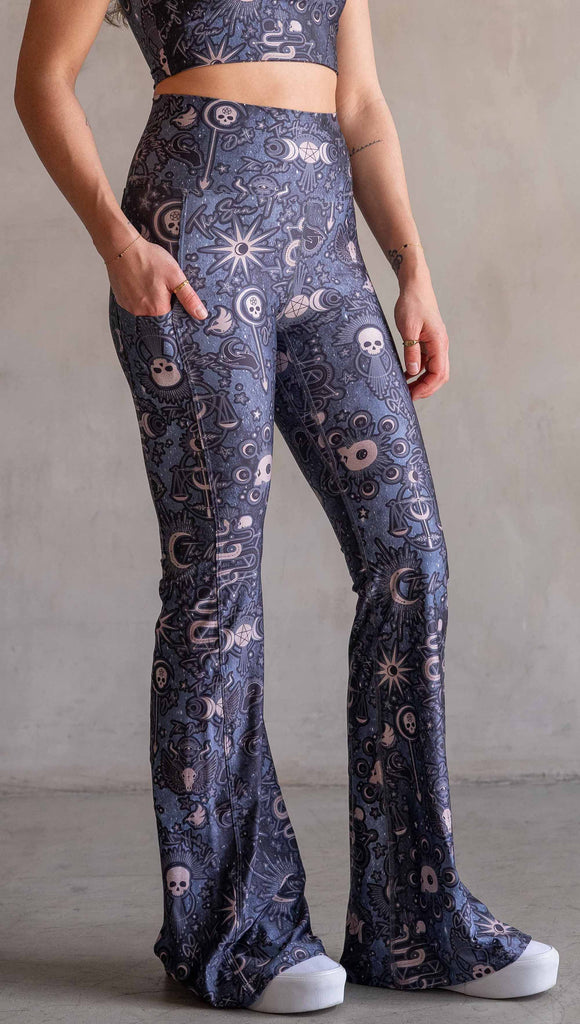 Model wearing WERKSHOP Tarot Bell Bottoms. They are high waist and feature pockets on both legs. The blue/tarot artwork has skulls, snakes, moons and the names of multiple popular tarot cards like "Strength, Lovers, Death and The Hanged Man”. Our model is 5’6” and this images shows her wearing the bells barefoot. The hem of the pants are hitting the floor.