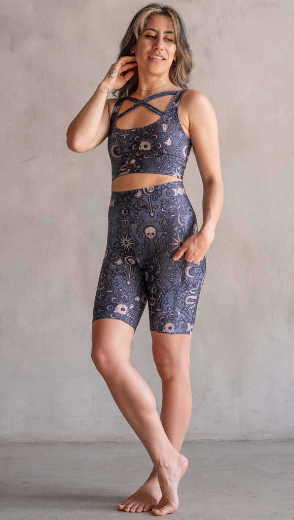 Model wearing WERKSHOP Tarot Bicycle Length Shorts. They are high waist and feature pockets on both legs. The blue/tarot artwork has skulls, snakes, moons and the names of multiple popular tarot cards like "Strength, Lovers, Death and The Hanged Man”. The length of the shorts hit in the mid thigh, above the knee.