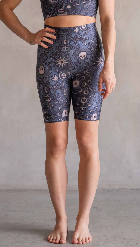 Model wearing WERKSHOP Tarot Bicycle Length Shorts. They are high waist and feature pockets on both legs. The blue/tarot artwork has skulls, snakes, moons and the names of multiple popular tarot cards like "Strength, Lovers, Death and The Hanged Man”. The length of the shorts hit in the mid thigh, above the knee.