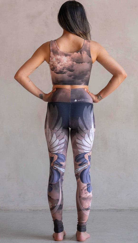 Full body back view of model wearing WERKSHOP Valkyrie Athleisure Leggings. featuring original artwork by Chriztina Marie. featuring a Valkyrie warrior from Norse Mythology flying through a dark sky holding a shield and swords. Her hair is in braids and she is wearing armor and a viking helmet. The colors are warm and the background also features a crescent moon and stars.