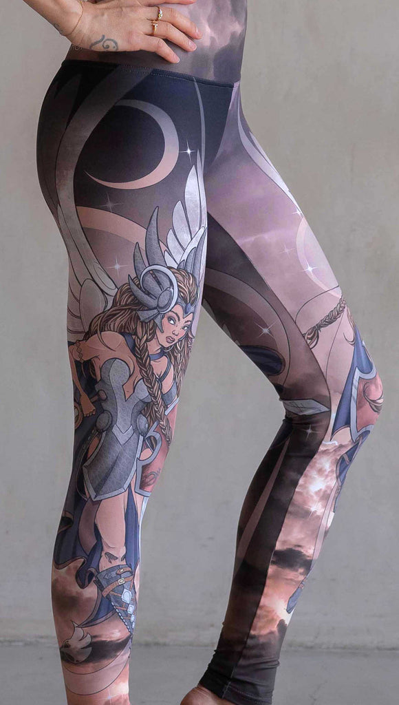 Zoomed in view of model wearing WERKSHOP Valkyrie Athleisure Leggings. featuring original artwork by Chriztina Marie. featuring a Valkyrie warrior from Norse Mythology flying through a dark sky holding a shield and swords. Her hair is in braids and she is wearing armor and a viking helmet. The colors are warm and the background also features a crescent moon and stars.
