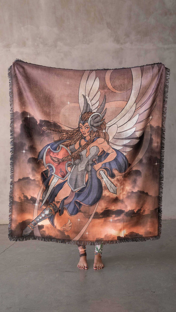 Decorative Chenille Tapestry printed with limited edition artwork by our Female Founder, Chriztina Marie. The artwork features a valyrkie warrior flying through a stormy sky holding a sword and shield. The colours are rich and warm with peach/orange tones and some pops of blue. This image shows a girl holding the tapestry up over her head to show off the artwork.