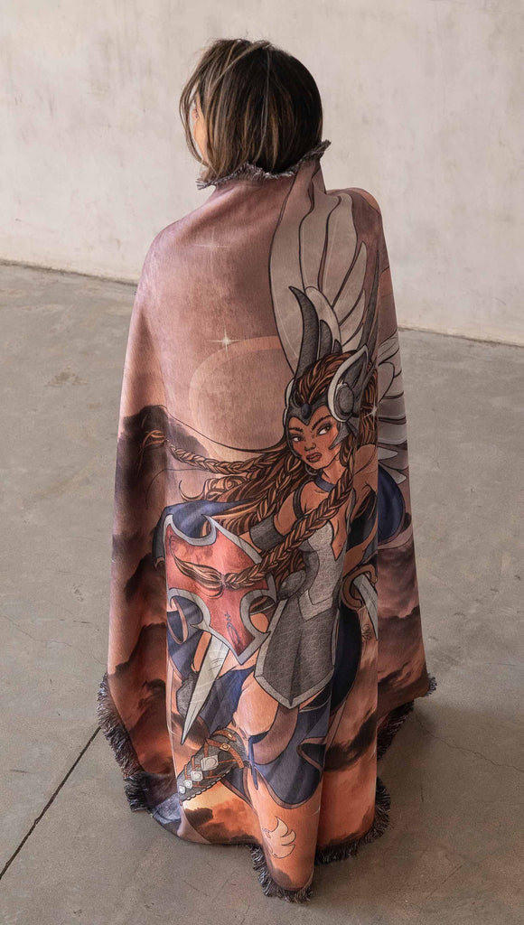 Decorative Chenille Tapestry printed with limited edition artwork by our Female Founder, Chriztina Marie. The artwork features a valyrkie warrior flying through a stormy sky holding a sword and shield. The colours are rich and warm with peach/orange tones and some pops of blue. This image shows a girl with the tapestry draped around her shouldeers like a cape.