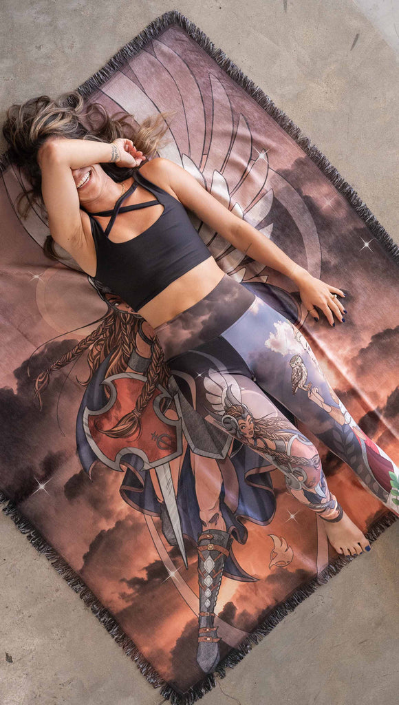 Decorative Chenille Tapestry printed with limited edition artwork by our Female Founder, Chriztina Marie. The artwork features a valyrkie warrior flying through a stormy sky holding a sword and shield. The colours are rich and warm with peach/orange tones and some pops of blue. This image shows a girl (Sam) laying on the tapestry and laughing.