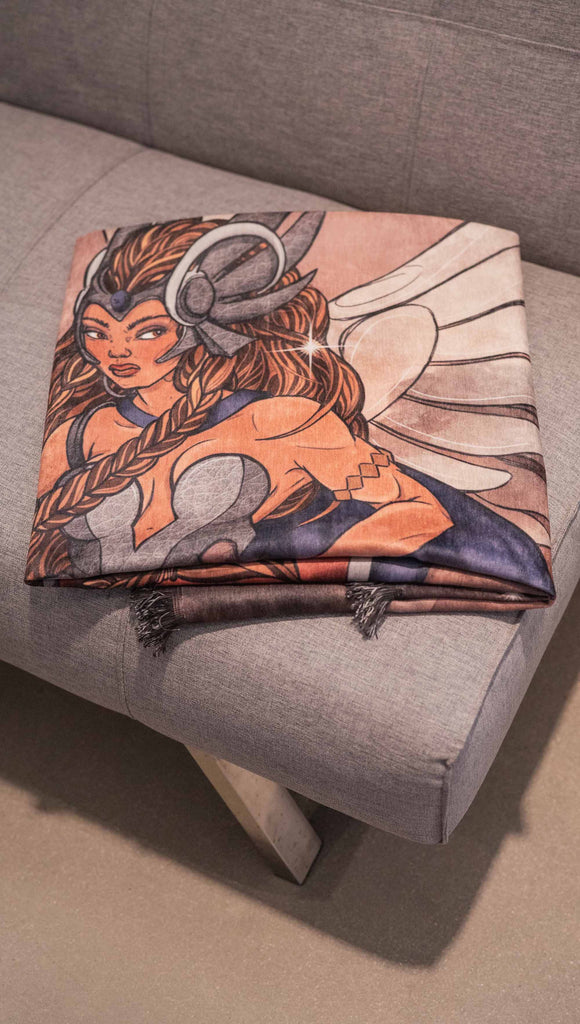 Decorative Chenille Tapestry printed with limited edition artwork by our Female Founder, Chriztina Marie. The artwork features a valyrkie warrior flying through a stormy sky holding a sword and shield. The colours are rich and warm with peach/orange tones and some pops of blue. This image shows the tapestry folded neatly on a couch.