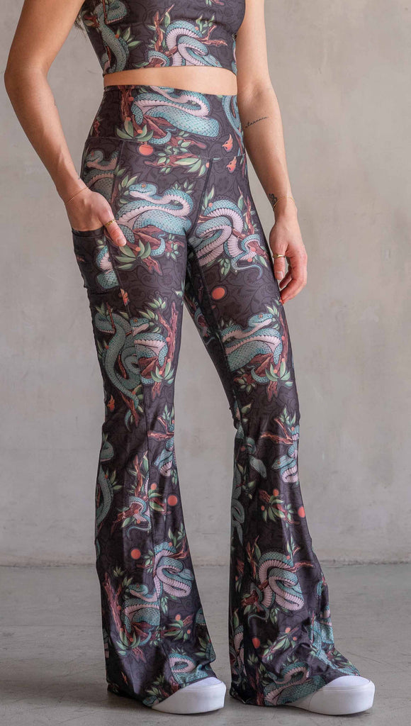 Model wearing WERKSHOP Pit Viper Featherlight Bells. The artwork on the leggings have clusters of teal blue pit viper snakes intertwined on tree branches over a taupe/brown background. The featherlight bells have pockets.